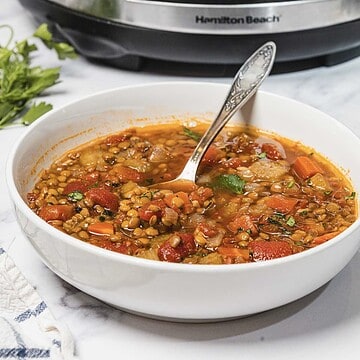 A bowl of Slow Cooker Lentil Soup with vegetables and a spoon sits on a white countertop near an Instant Pot.