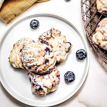 Iced blueberry cookies on a white plate with fresh blueberries and air fryer blueberry donut holes beside them, seen from above.