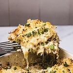 A fork lifting a cheesy, golden baked Tuna Casserole with peas and herbs from a baking dish.