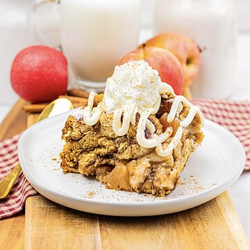 A slice of slow cooker apple pie cinnamon roll casserole is topped with whipped cream and icing on a plate, with apples and milk in the background.