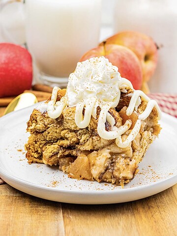 A slice of slow cooker apple pie cinnamon roll casserole is topped with whipped cream and icing on a plate, with apples and milk in the background.