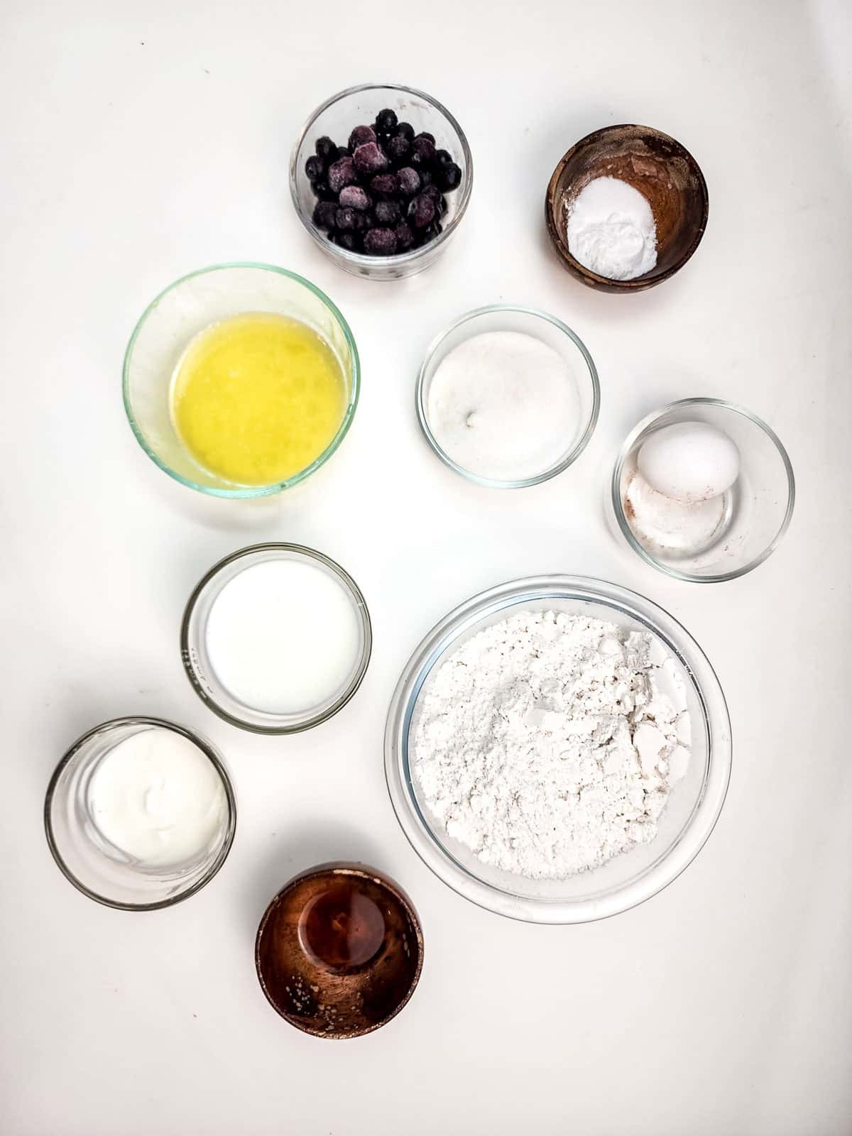 Nine bowls with ingredients for baking: flour, blueberries, egg, sugar, milk, melted butter, yogurt, cocoa, and baking powder—perfect for making delicious Blueberry Donut Holes in your Air Fryer.