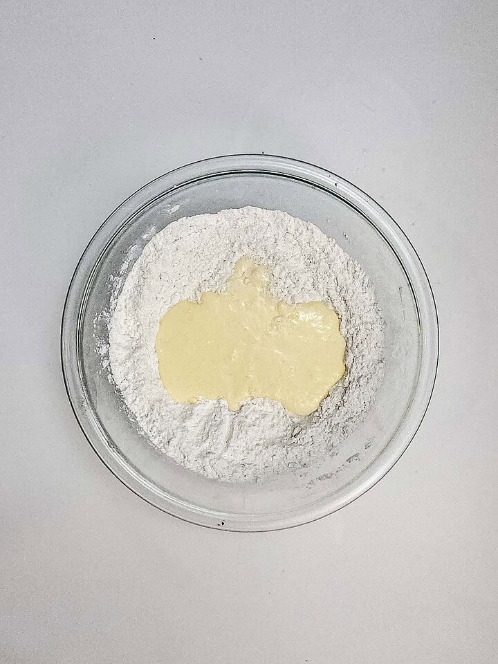 Clear glass bowl with flour and a pool of liquid batter in the center, perfect for making Air Fryer Blueberry Donut Holes, on a white surface.