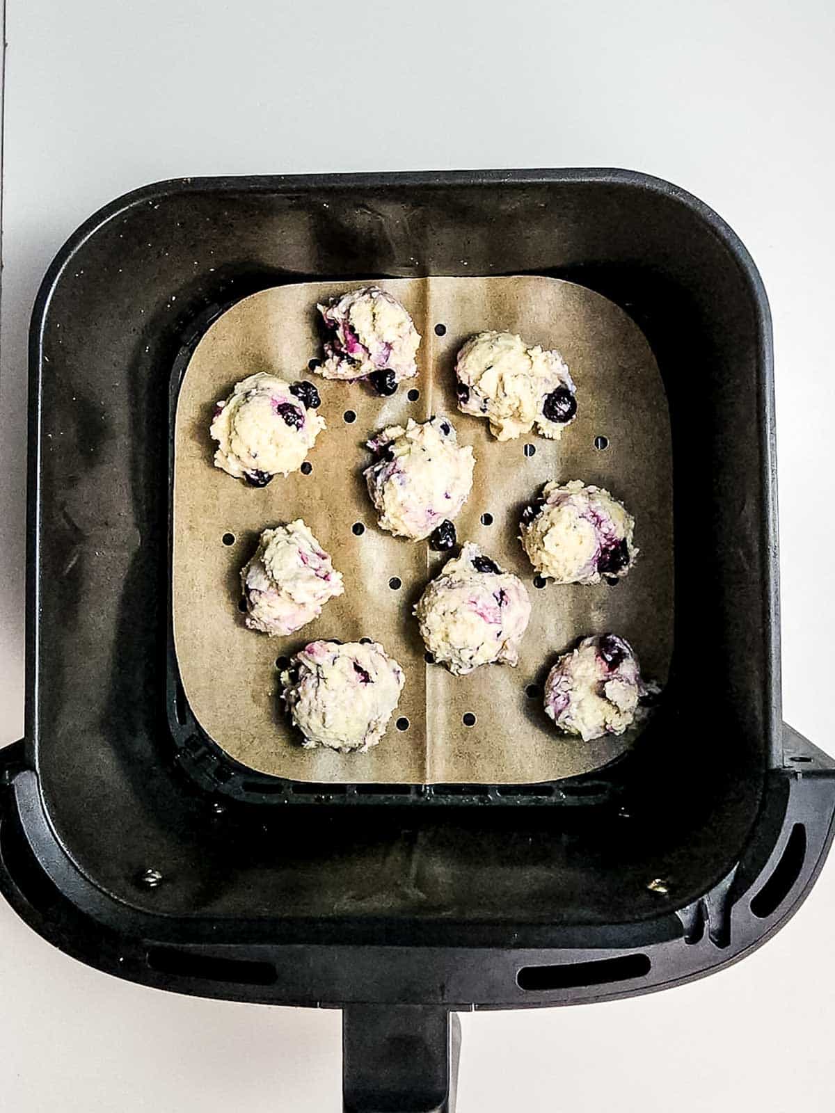 Nine unbaked blueberry donut holes rest on parchment paper in an air fryer basket, ready to become delicious treats.