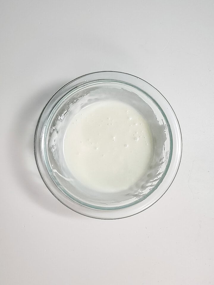 A glass bowl with a small amount of white donut hole batter rests on a white surface, ready to make delicious air fryer treats.