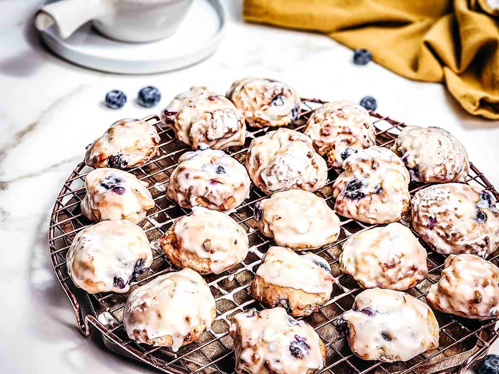 Glazed blueberry cookies cooling on a round wire rack, with scattered blueberries and a yellow cloth nearby—these treats look like blueberry donut holes fresh from the air fryer.