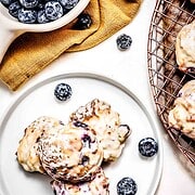 Three glazed blueberry scones on a white plate, surrounded by fresh blueberries and a bowl nearby—perfect for pairing with homemade Air Fryer Blueberry Donut Holes.