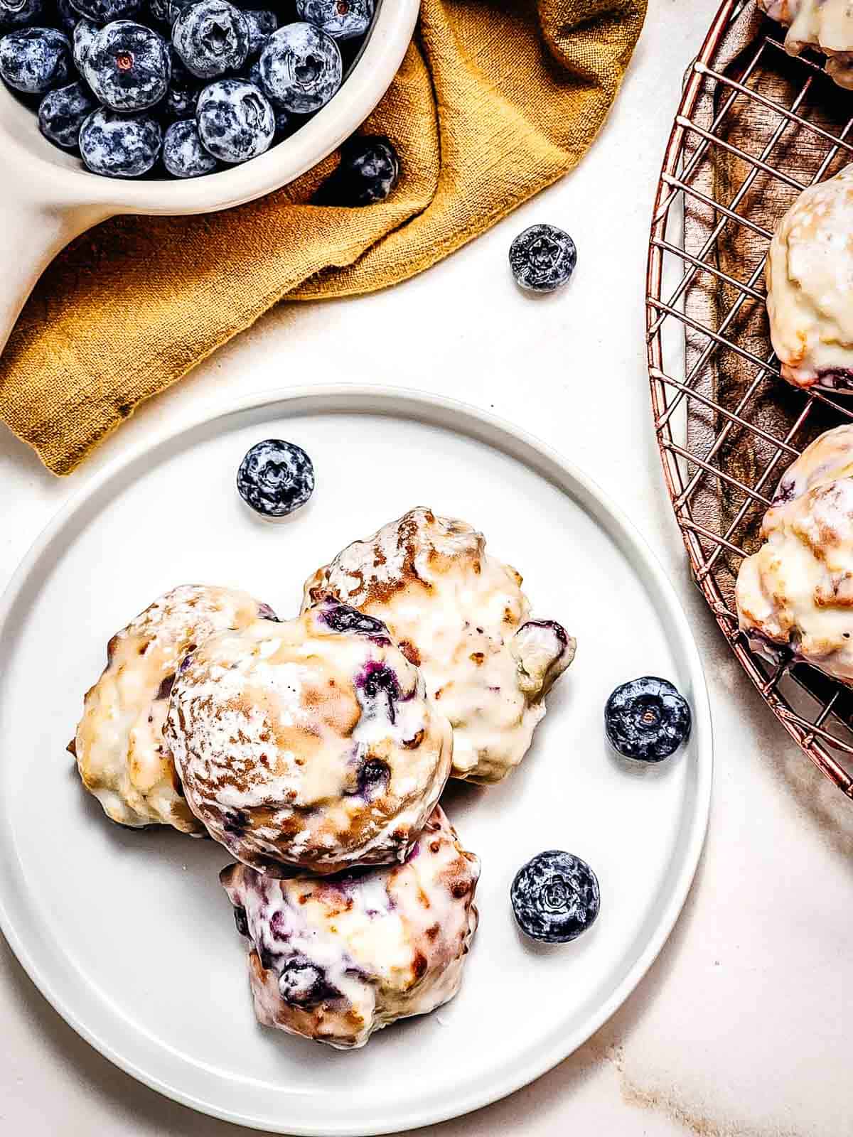 Three glazed blueberry scones on a white plate, surrounded by fresh blueberries and a bowl nearby—perfect for pairing with homemade Air Fryer Blueberry Donut Holes.