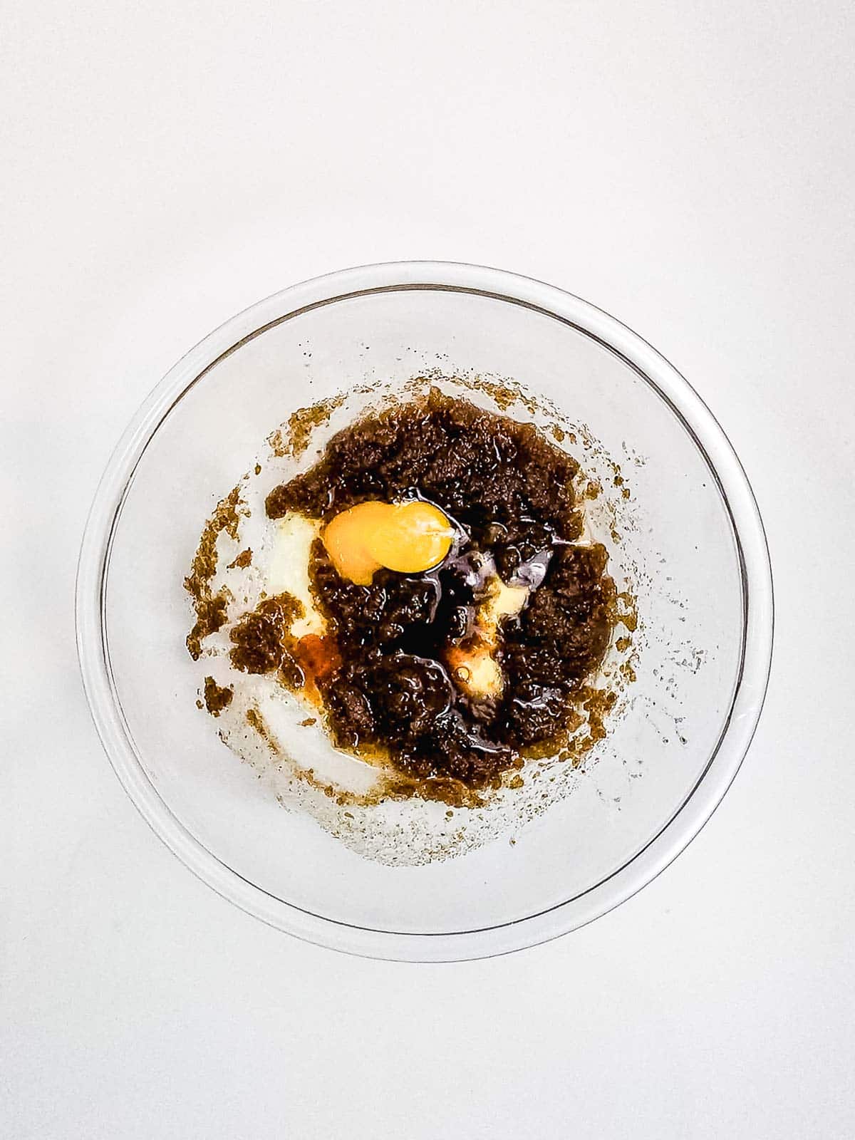A glass bowl with brown sugar, butter, and an egg on a white surface, ready for mixing delicious Chocolate Chunk Cookies.