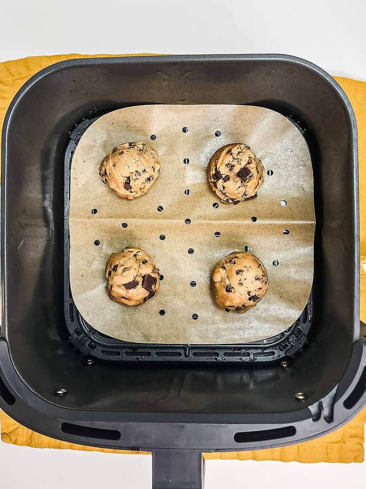 Four chocolate chunk cookie dough balls on parchment paper inside an air fryer basket, ready to become delicious Air Fryer Cookies.