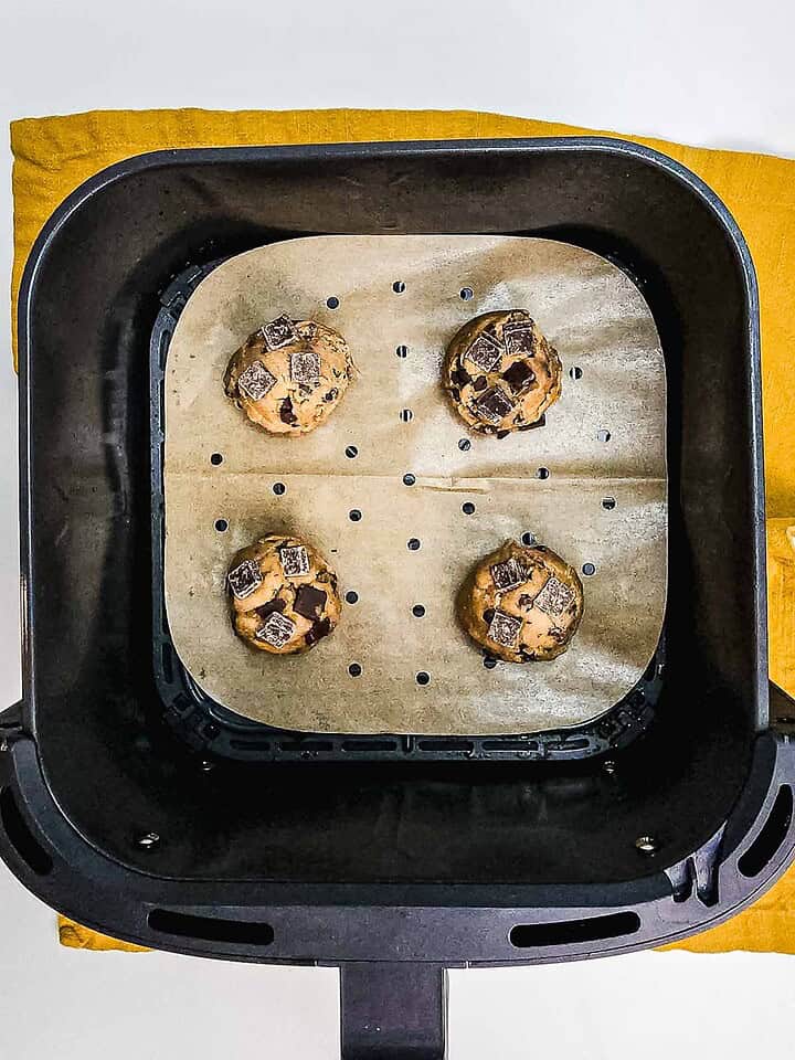 Four unbaked chocolate chunk cookies on parchment paper in an air fryer basket, set atop a yellow cloth.