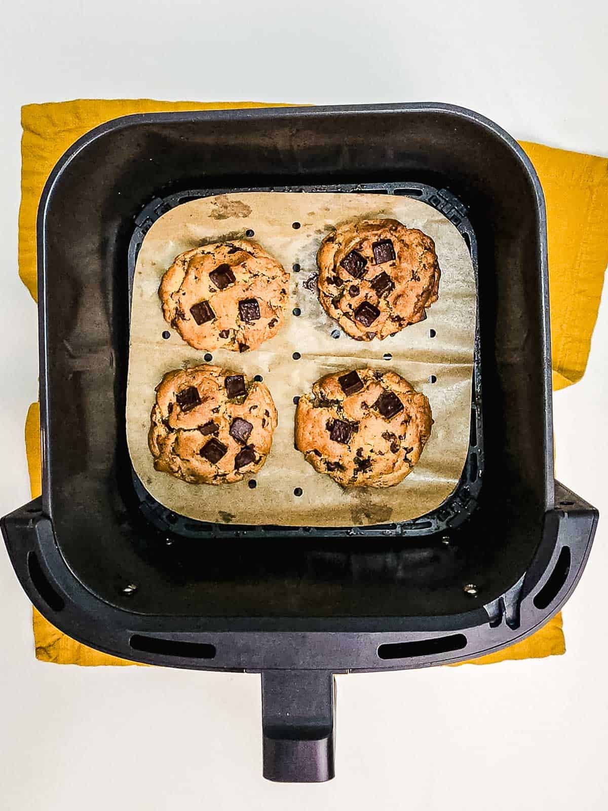 Four chocolate chunk cookies on parchment paper in an air fryer basket, viewed from above, showcase the ease of making delicious treats with your Air Fryer.