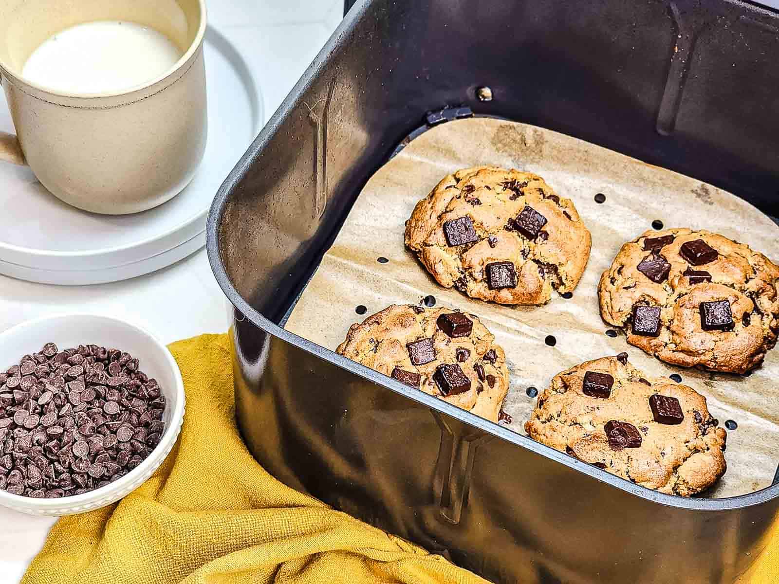 Four Chocolate Chunk Cookies in an air fryer basket, with milk and chocolate chips nearby on a yellow cloth.
