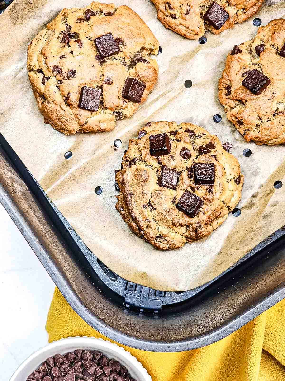 Chocolate chunk cookies with gooey chips rest on parchment in an air fryer basket, with a bowl of extra chocolate chips nearby.