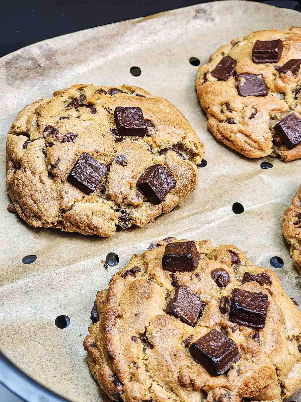 Three large chocolate chunk cookies with gooey chocolate on top, freshly baked in the air fryer and resting on parchment paper.