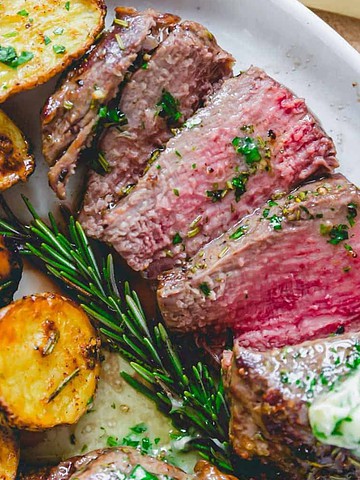 Sliced steak with herb butter, roasted potatoes, and rosemary on a white plate.