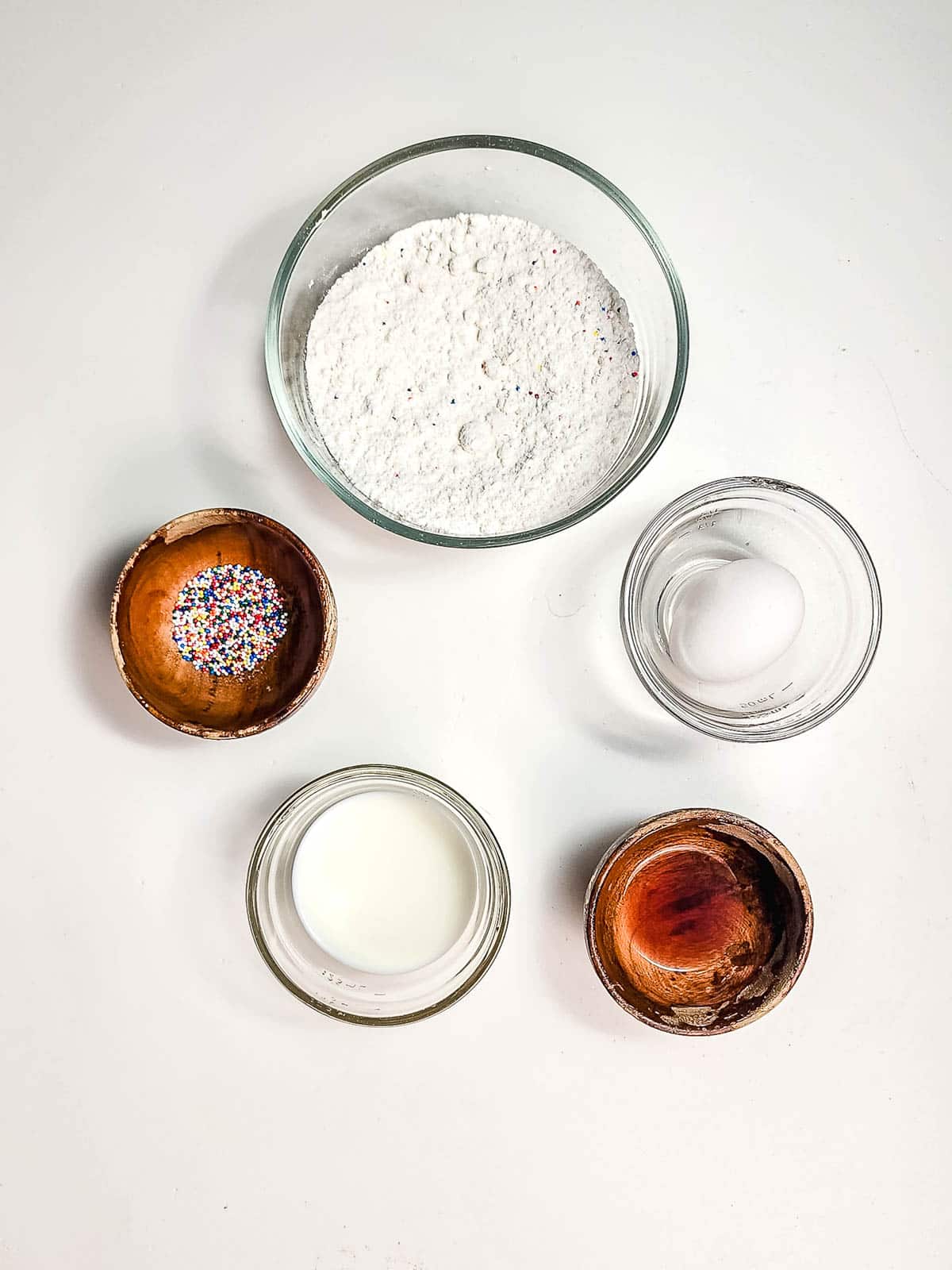 Five bowls with cake mix, an egg, milk, rainbow sprinkles, and oil on a white surface—perfect for making delicious Funnel Cake Bites with this easy Air Fryer recipe.