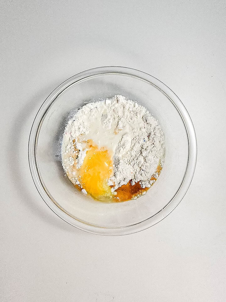A glass bowl with flour, an egg, milk, and vanilla ready to be mixed for a delicious Funnel Cake Bites recipe on a white surface.
