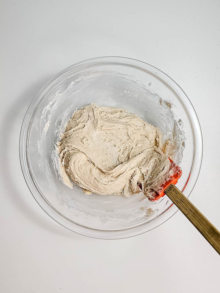 A glass bowl with thick funnel cake dough being mixed by a red spatula on a white surface, perfect for your next air fryer recipe.