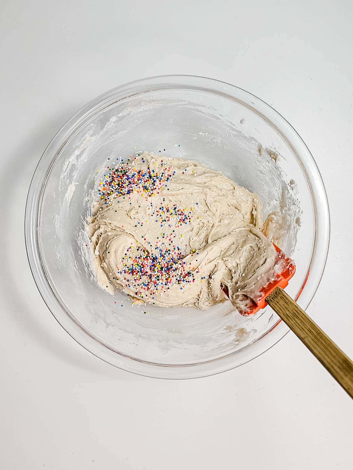 A glass bowl of cake batter with rainbow sprinkles and a red spatula on a white surface, perfect for trying a new funnel cake bites recipe.