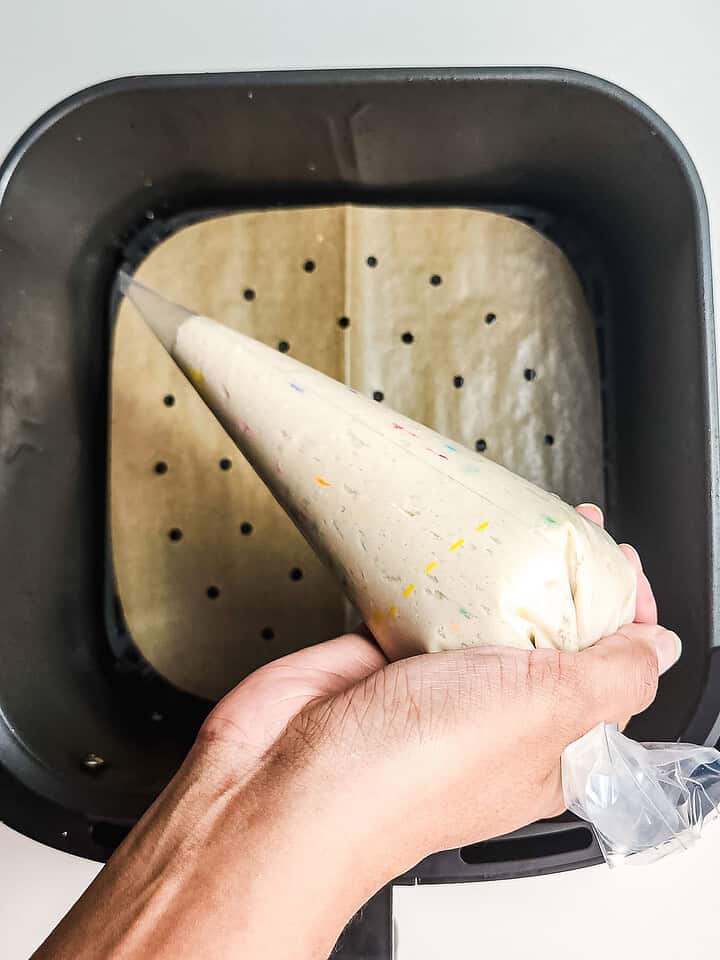 Hand holding a piping bag with batter over parchment paper in an air fryer basket, perfect for making funnel cake bites with this easy recipe.