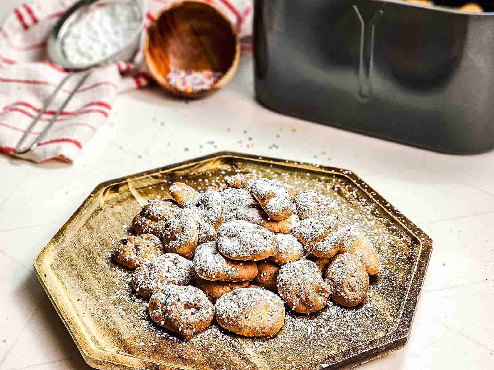 Golden Funnel Cake Bites dusted with powdered sugar on a tray, with baking tools and sprinkles in the background—a perfect air fryer recipe for sweet cravings.