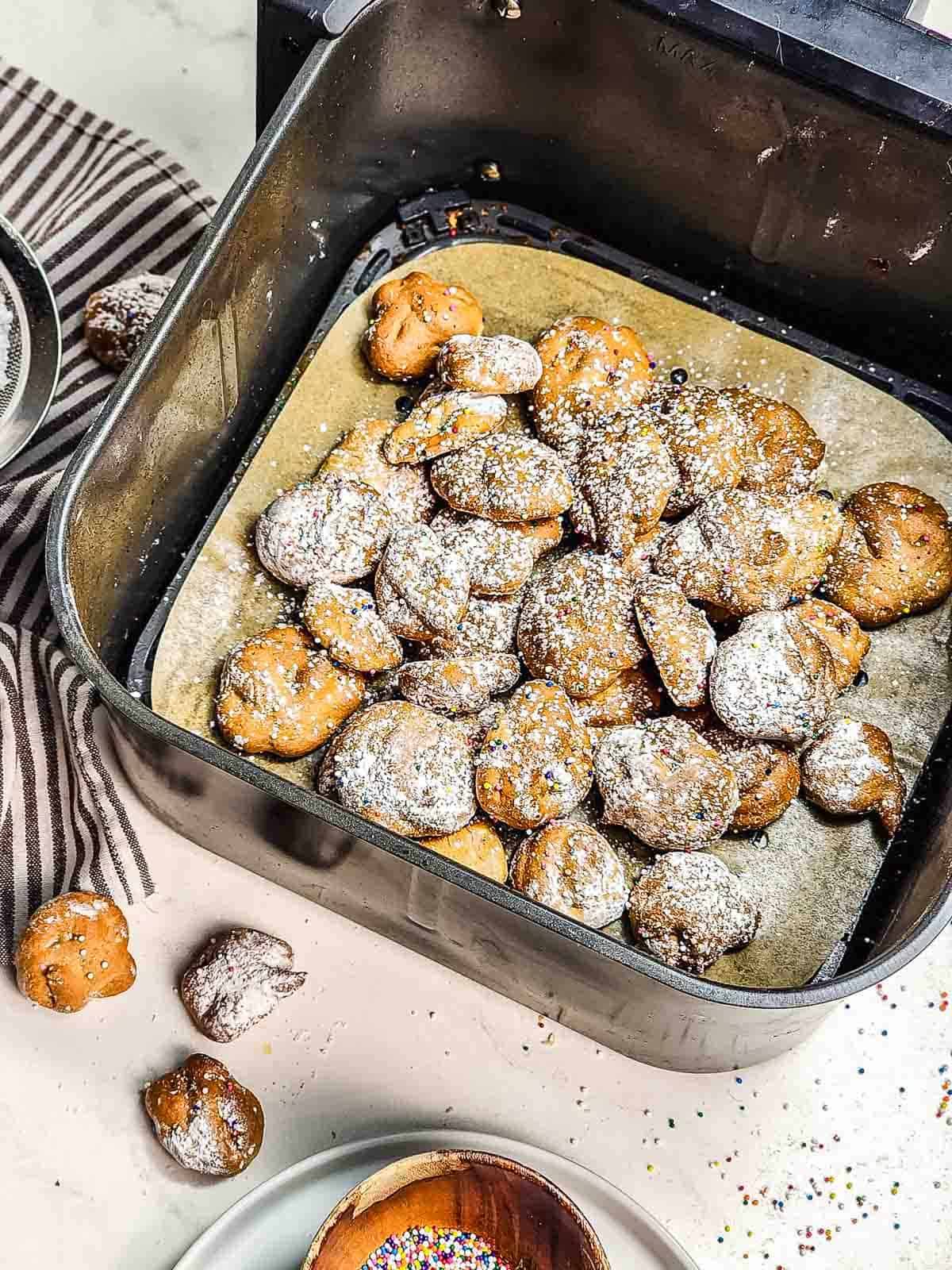 Funnel Cake Bites and powdered sugar donut holes in an air fryer basket lined with parchment paper, with sprinkles nearby—an irresistible recipe for sweet snacking!.