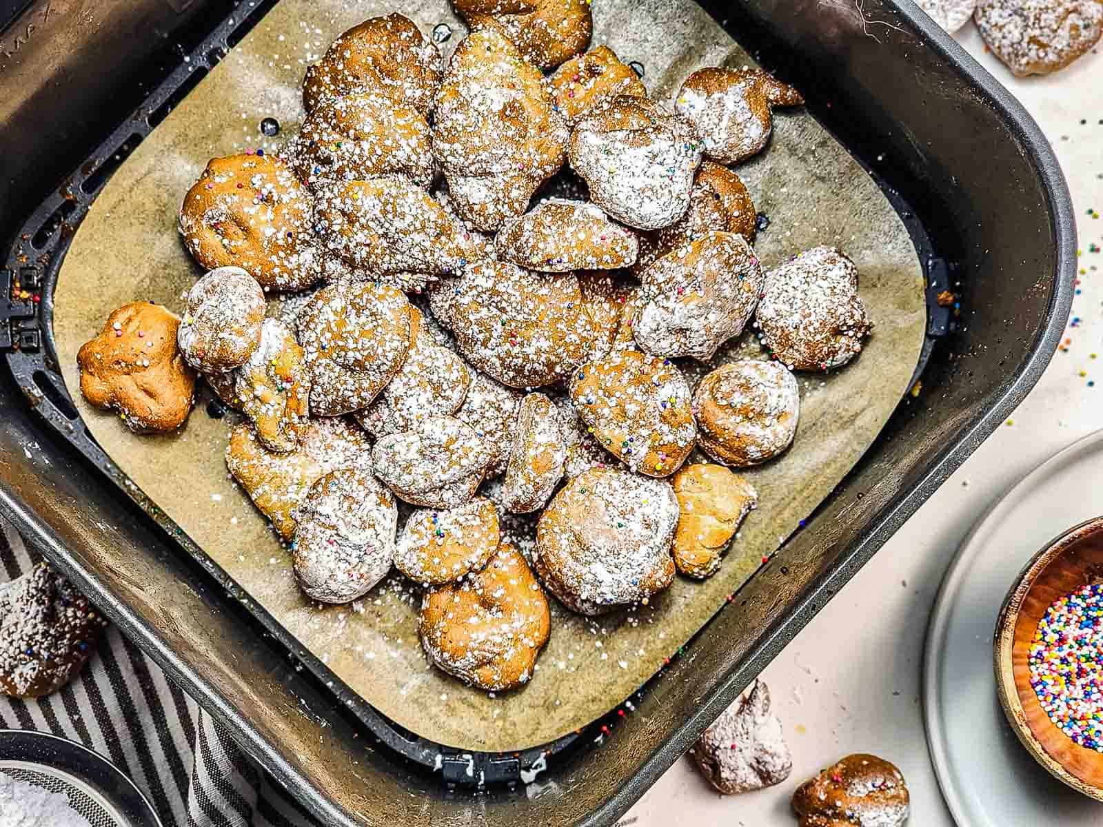Air fryer basket with golden brown Funnel Cake Bites dusted with powdered sugar on parchment paper—an irresistible recipe for sweet, crisp treats.