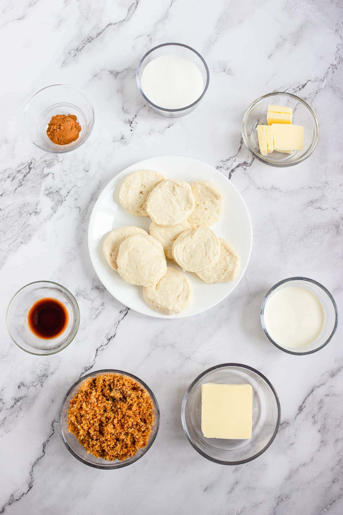 Ingredients for baking on a marble surface, including biscuits, butter, milk, sugar, vanilla, and cinnamon.