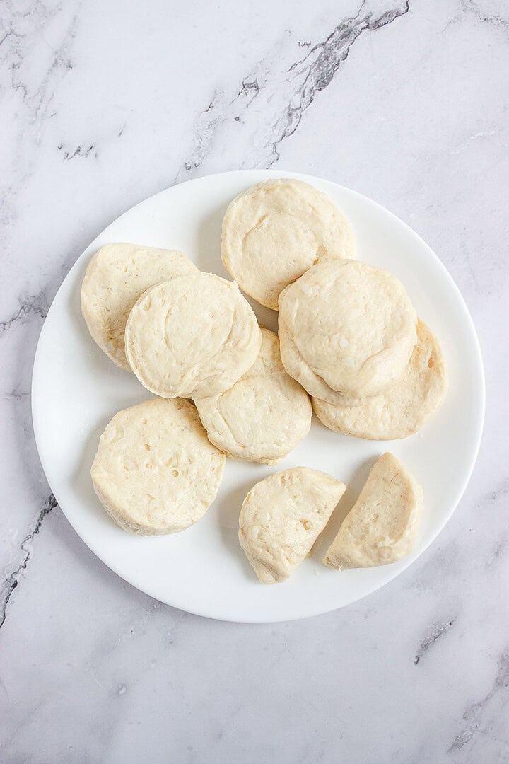 A white plate with several uncooked biscuit dough rounds on a marble surface.