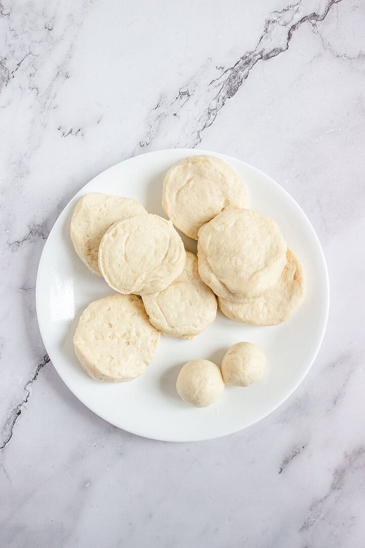 A white plate with raw, round biscuit dough pieces and two dough balls on a marble surface.