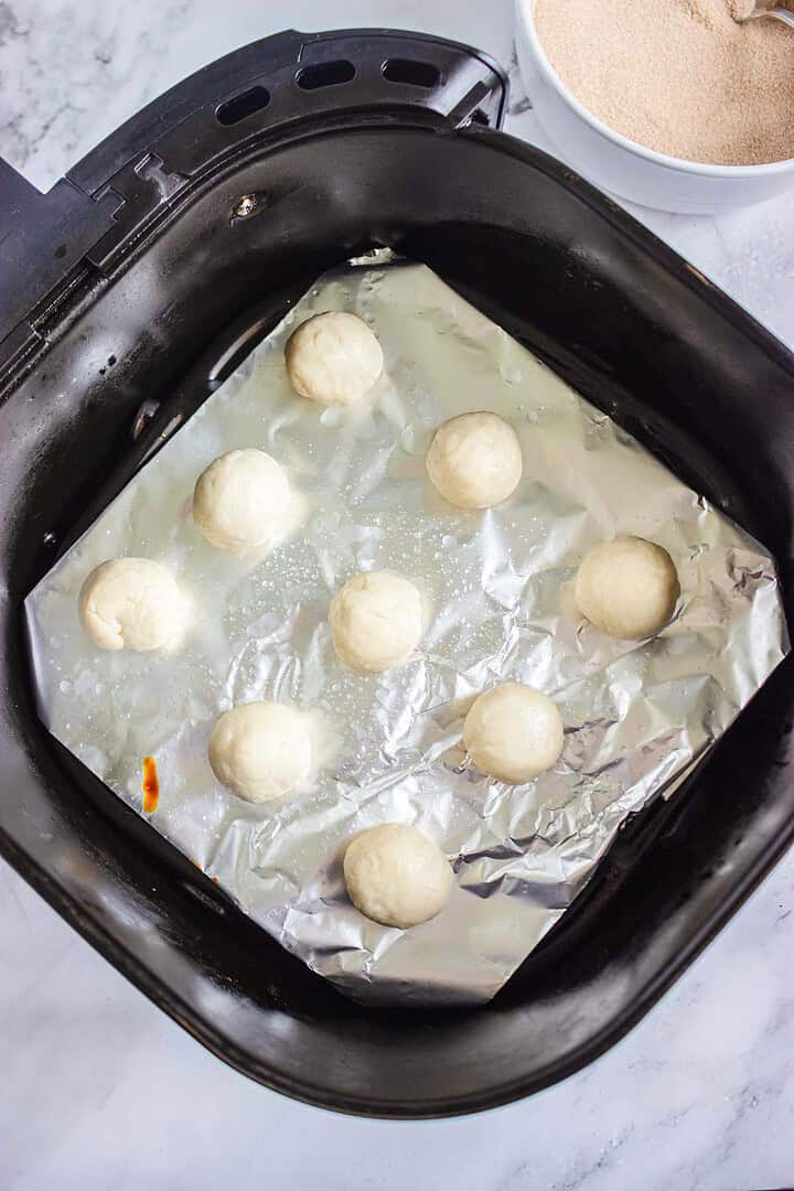 Eight Churro Donut Holes rest on foil in an air fryer basket, with a bowl of breadcrumbs nearby&mdash;perfectly crisped and ready to dip in caramel sauce.