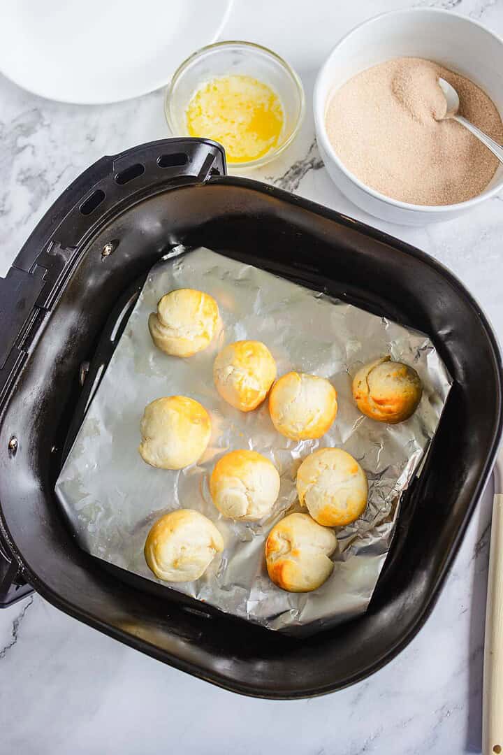 Nine churro donut holes baked in an air fryer basket on foil, with bowls of melted butter and cinnamon sugar nearby.