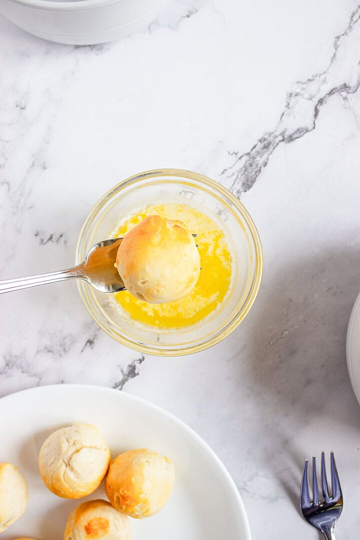 A spoon dips a bread roll into melted butter, with more rolls on a white plate nearby&mdash;perfect for pairing with churro donut holes or drizzling caramel sauce for an extra touch of sweetness.