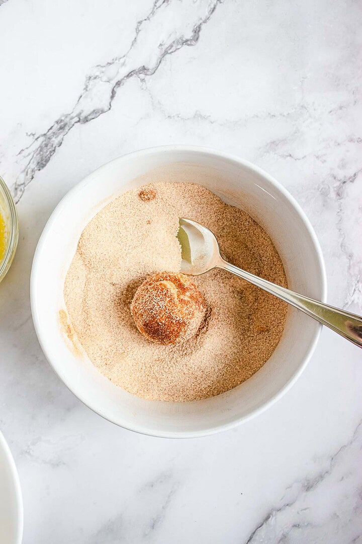A dough ball for Air Fryer Churro Donut Holes being coated in cinnamon sugar in a white bowl with a spoon on a marble surface, perfect for dipping in caramel sauce.