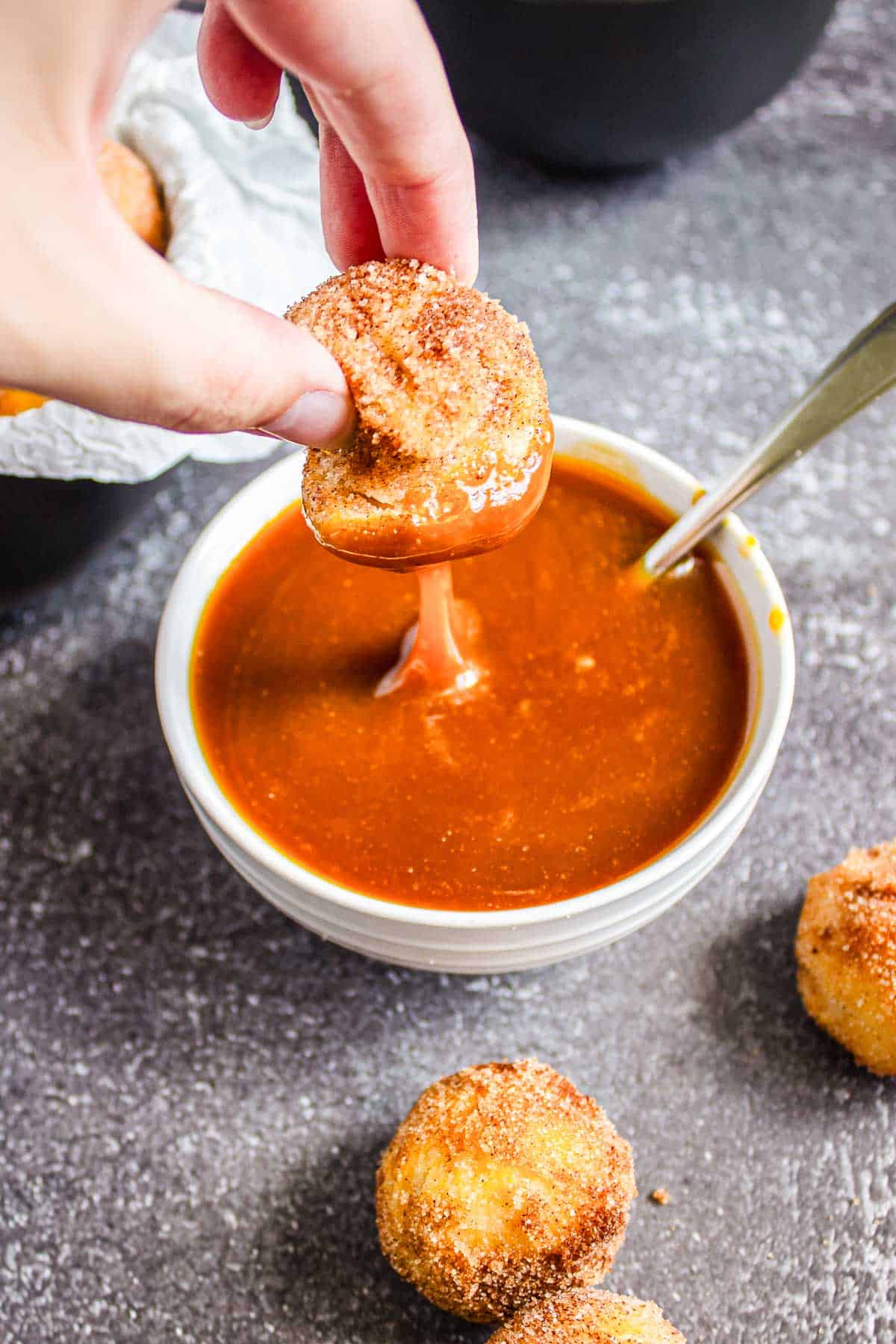 Hand dipping a churro donut hole into a bowl of caramel sauce, with more air fryer donut holes nearby.