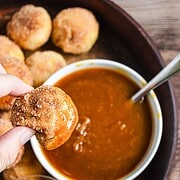 A hand holds a cinnamon sugar Air Fryer Churro Donut Hole dipped in caramel sauce, with more donut holes on a tray.