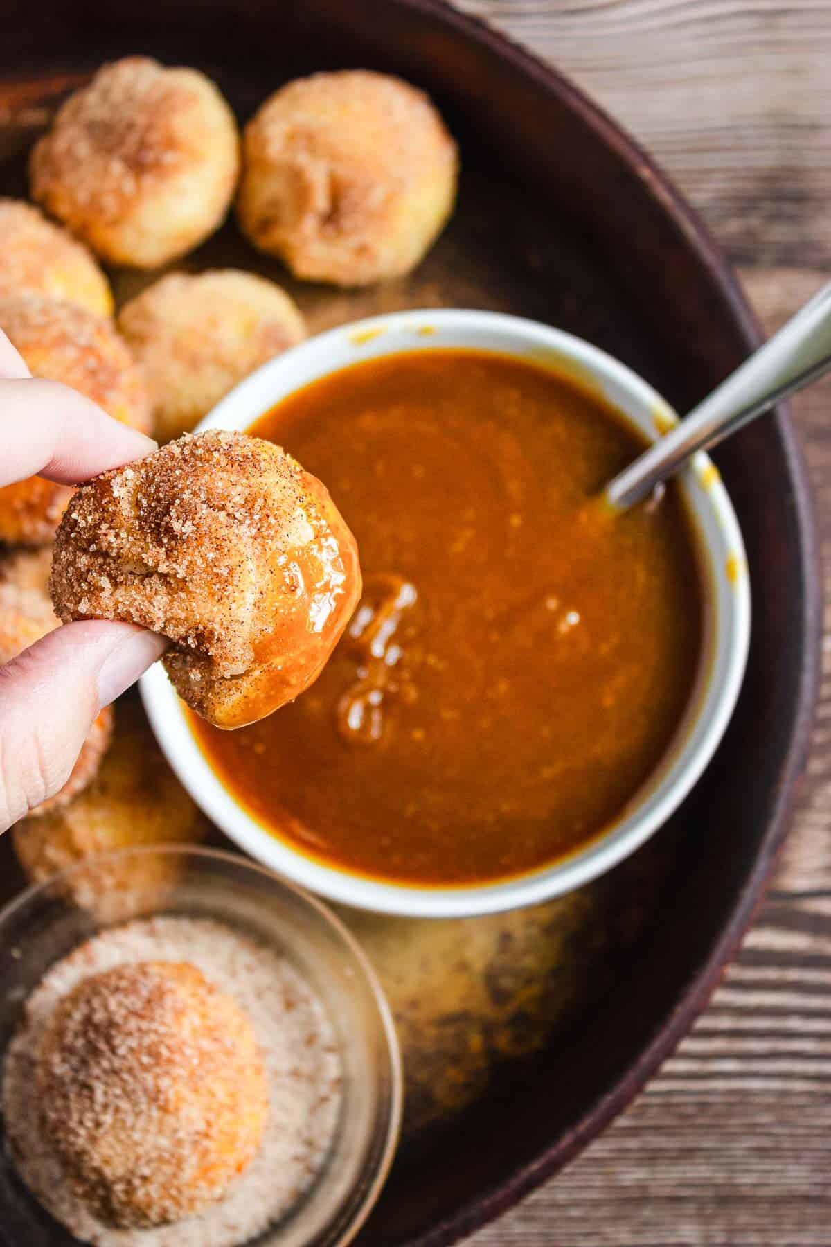 A hand holds a cinnamon sugar Air Fryer Churro Donut Hole dipped in caramel sauce, with more donut holes on a tray.