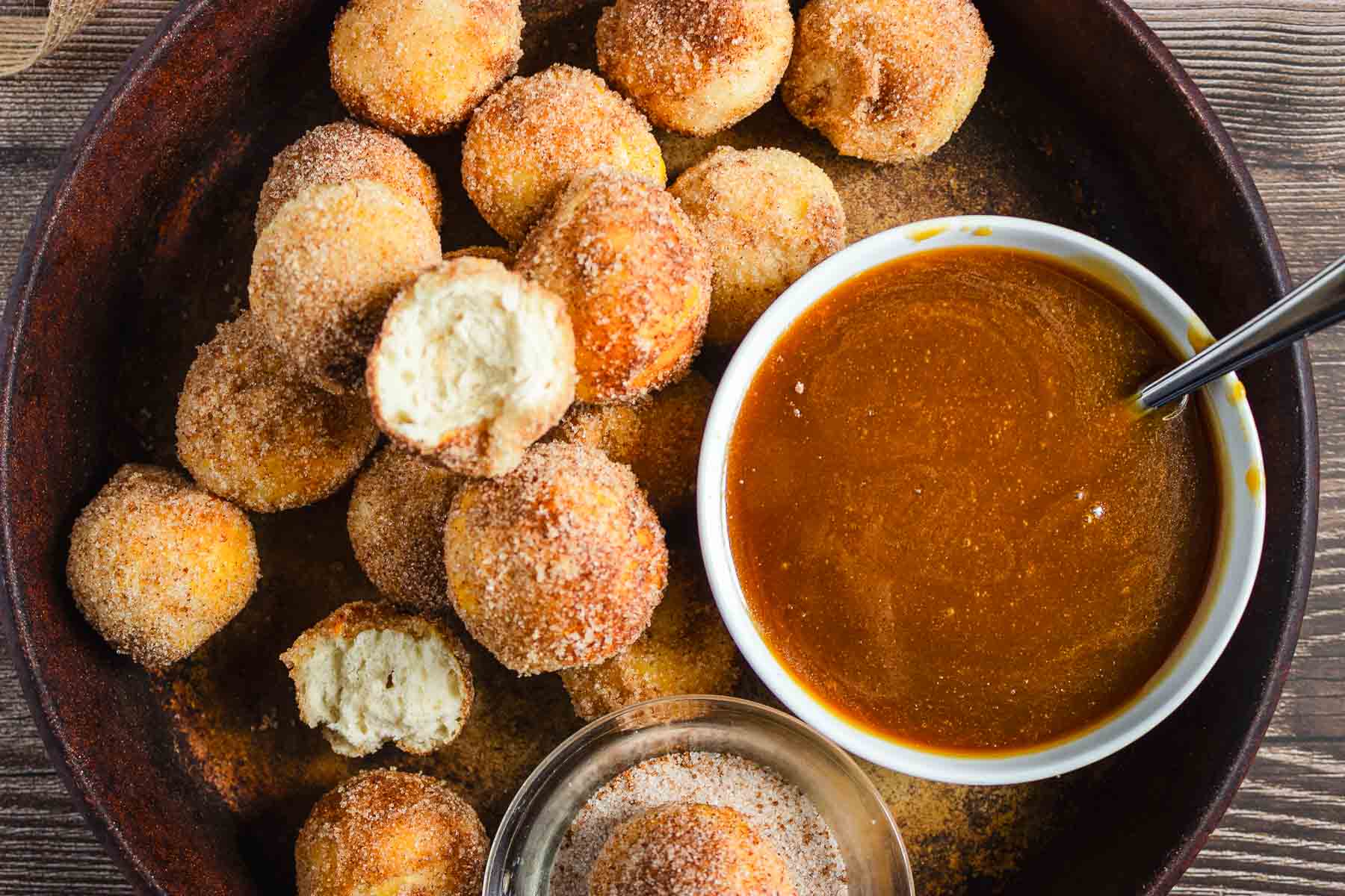 Round churro donut holes on a plate with cinnamon sugar, served alongside a bowl of rich caramel sauce and a spoon for dipping.