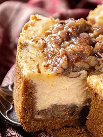 A close-up of a sliced cheesecake topped with chopped pecans and caramel, on a red patterned cloth.