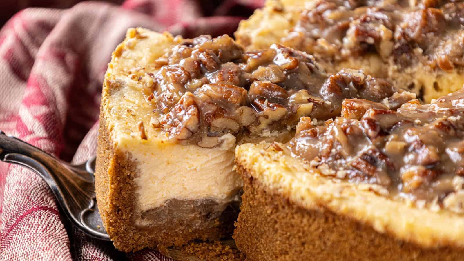 A close-up of a sliced cheesecake with a pecan topping on a graham cracker crust, placed on a red-striped cloth.
