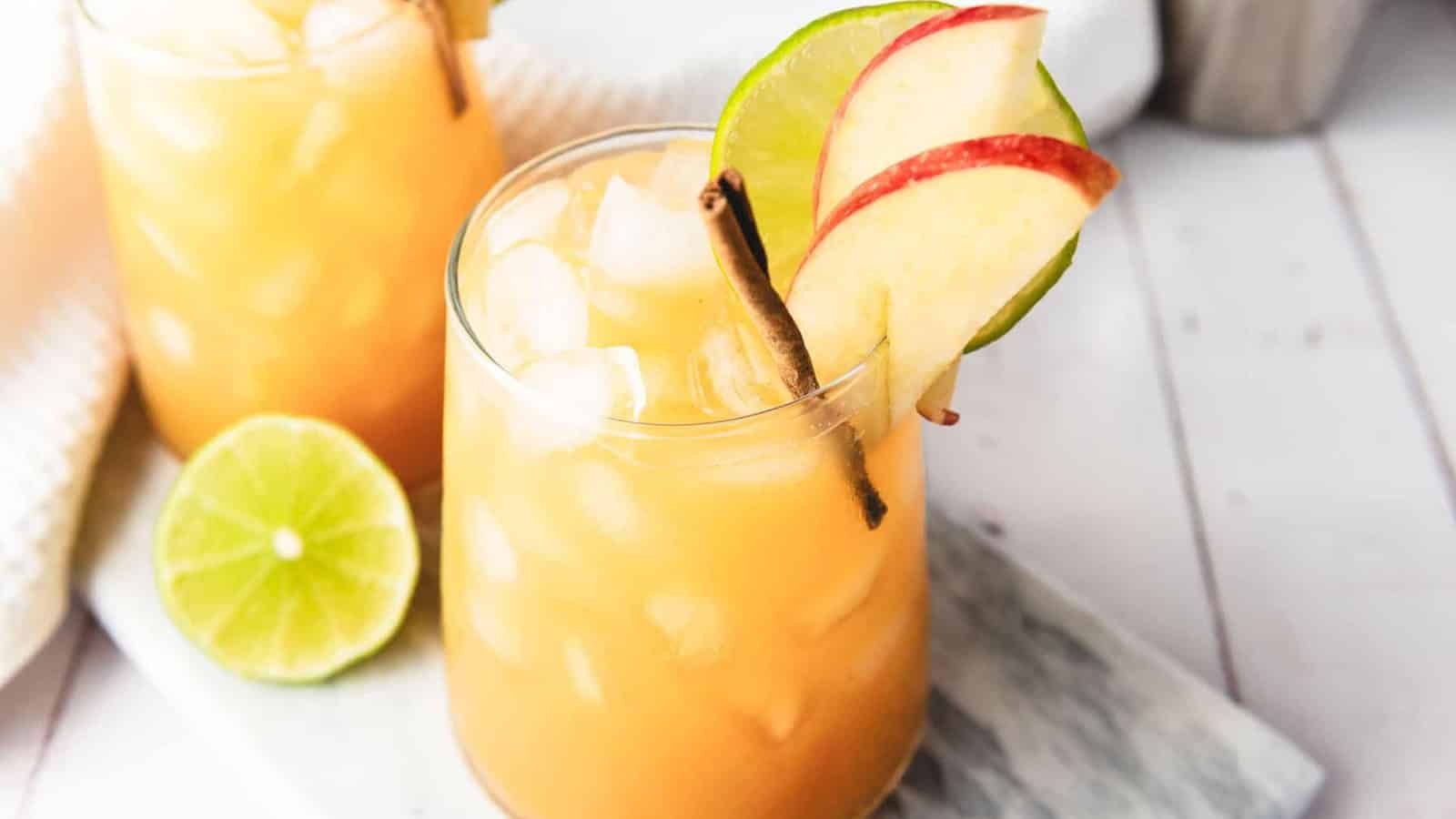Iced apple cider drink garnished with apple slices, a lime wheel, and a cinnamon stick on a marble tray.