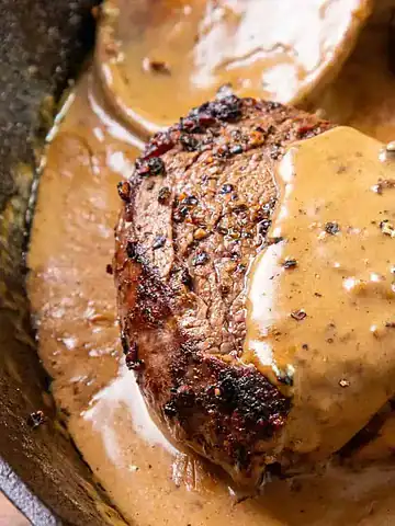 A seared steak topped with creamy peppercorn sauce in a skillet.