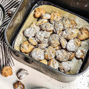 Air Fryer Funnel Cake Bites—small round fritters dusted with powdered sugar—rest in an air fryer basket lined with parchment paper in this easy recipe.