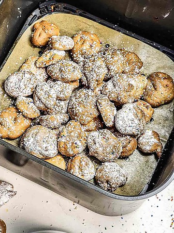 Air Fryer Funnel Cake Bites—small round fritters dusted with powdered sugar—rest in an air fryer basket lined with parchment paper in this easy recipe.