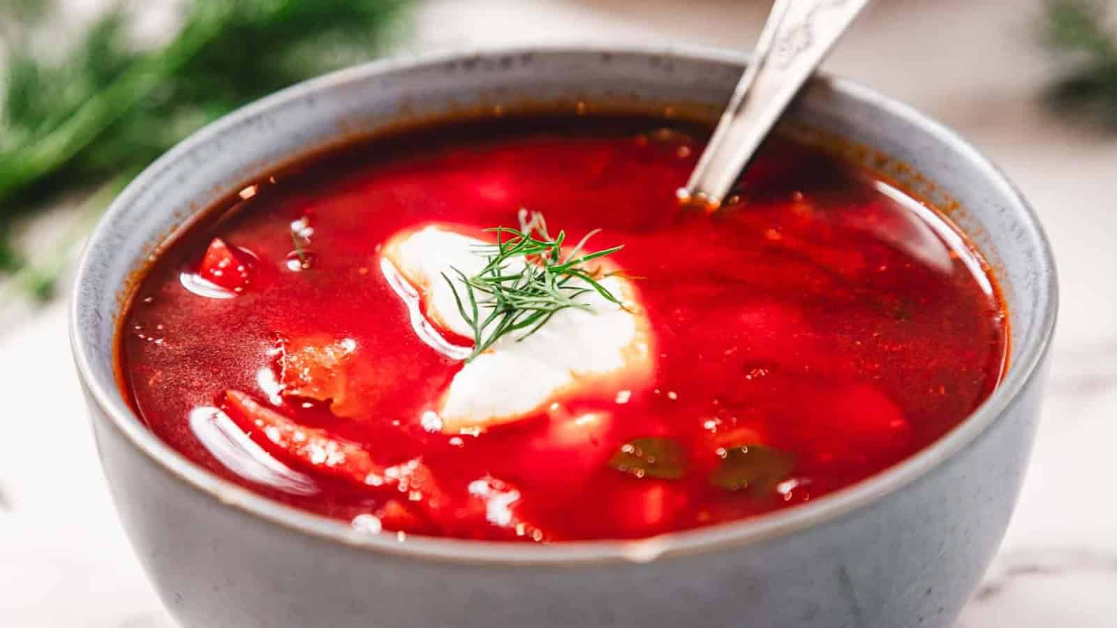 A bowl of borscht topped with sour cream and fresh dill, with a spoon in the bowl.