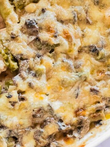 A baked casserole with melted cheese, chicken, mushrooms, and asparagus in a white dish with a serving spoon.