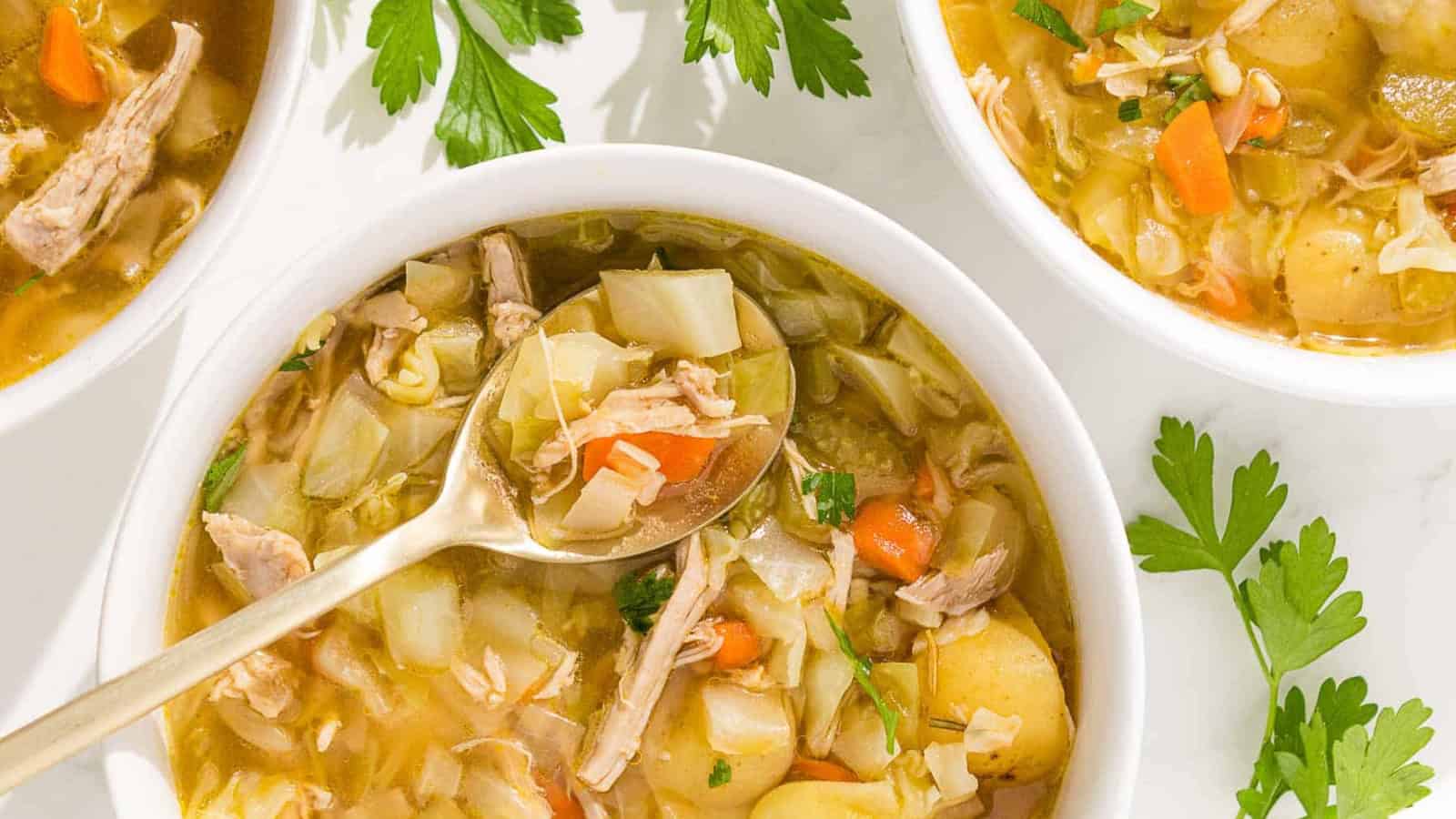 A bowl of chicken and cabbage soup is shown on a rustic wooden cutting board with parsley and a red linen.