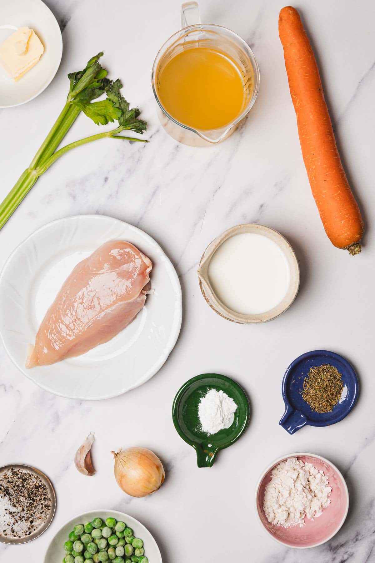 Ingredients for Chicken and Dumplings: raw chicken breast, carrot, celery, broth, cream, butter, onion, garlic, peas, spices, and flour arranged on a marble surface. Try this for your next Easy Chicken Recipes night!.