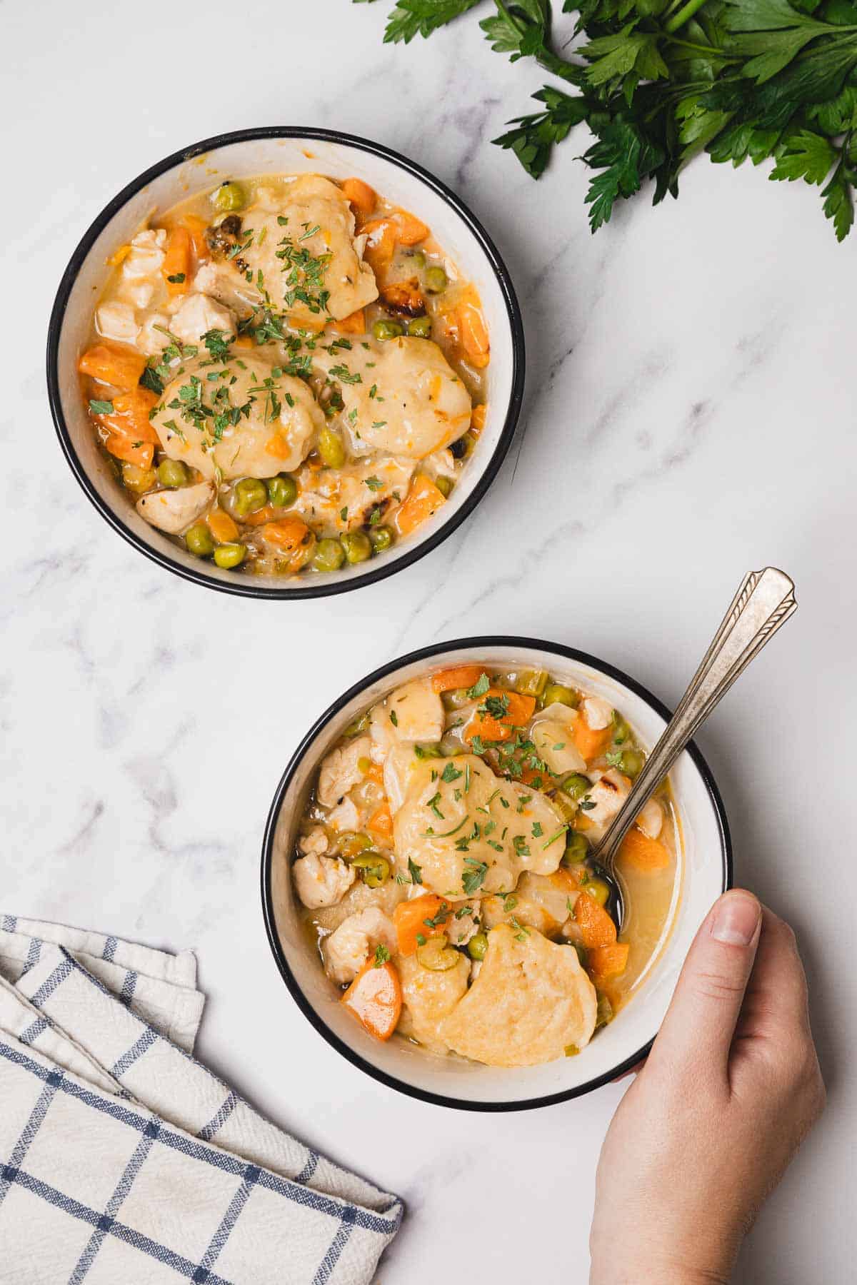 Two bowls of Chicken and Dumplings stew with carrots and peas, one held by a hand, on a white surface make for an easy skillet recipe perfect for cozy meals.